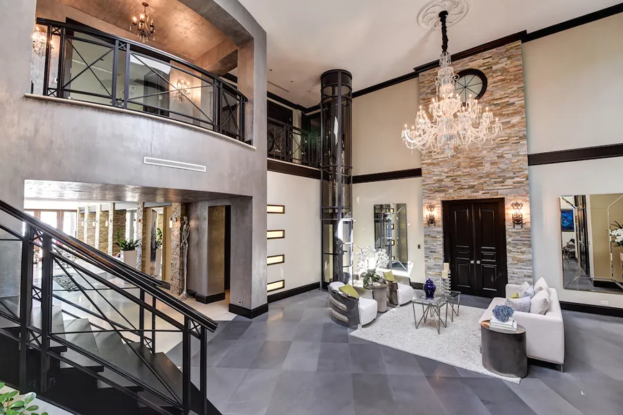 Ampio soggiorno moderno a doppia altezza con ascensore domestico panoramico, scala interna e zona living elegante, esempio di soluzioni per l’accessibilità verticale in casa